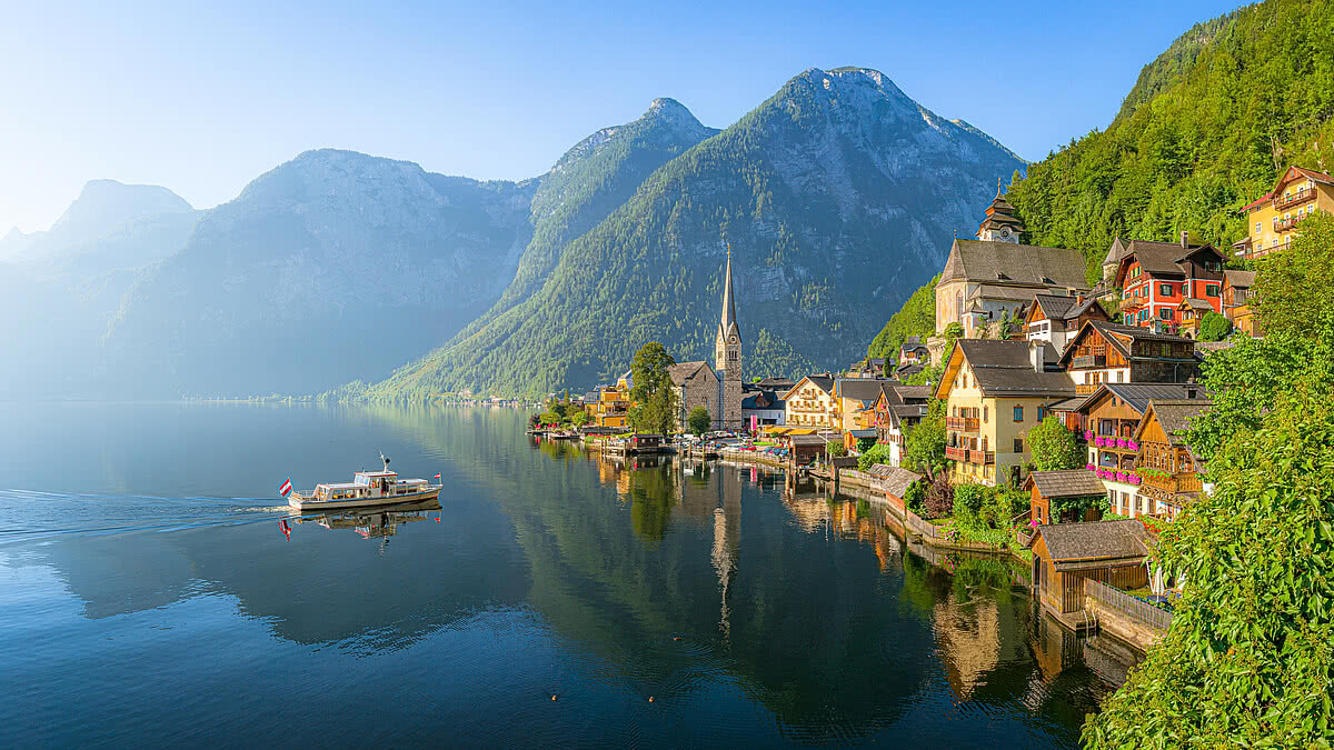 Classic panoramic view of idyllic Hallstatt lake in the Alps with famous old town with tourist ship in scenic golden morning light on a beautiful sunny day at sunrise in summer, Salzkammergut region, Austria